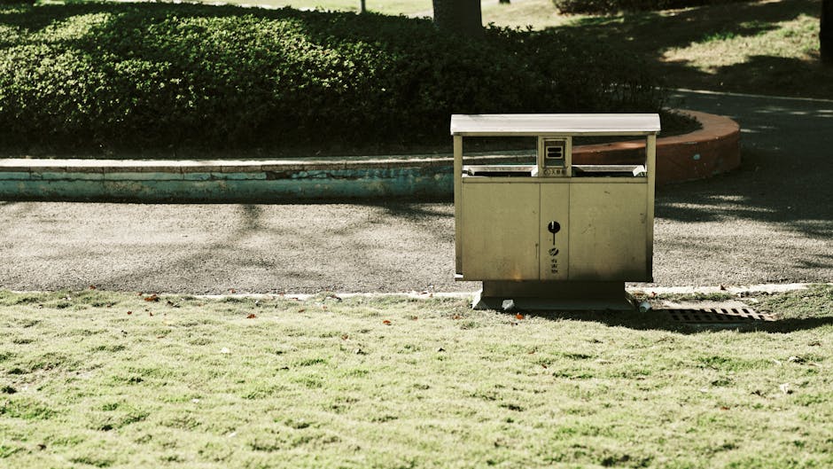 A small, metallic garden waste collection point situated on a grassy verge next to a paved pathway. The container has a simple, boxy design with a hinged lid at the top, possibly for depositing garden