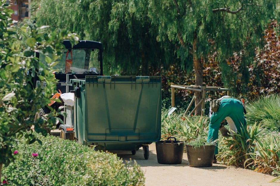 A small, metallic garden waste collection point situated on a grassy verge next to a paved pathway. The container has a simple, boxy design with a hinged lid at the top, possibly for depositing garden